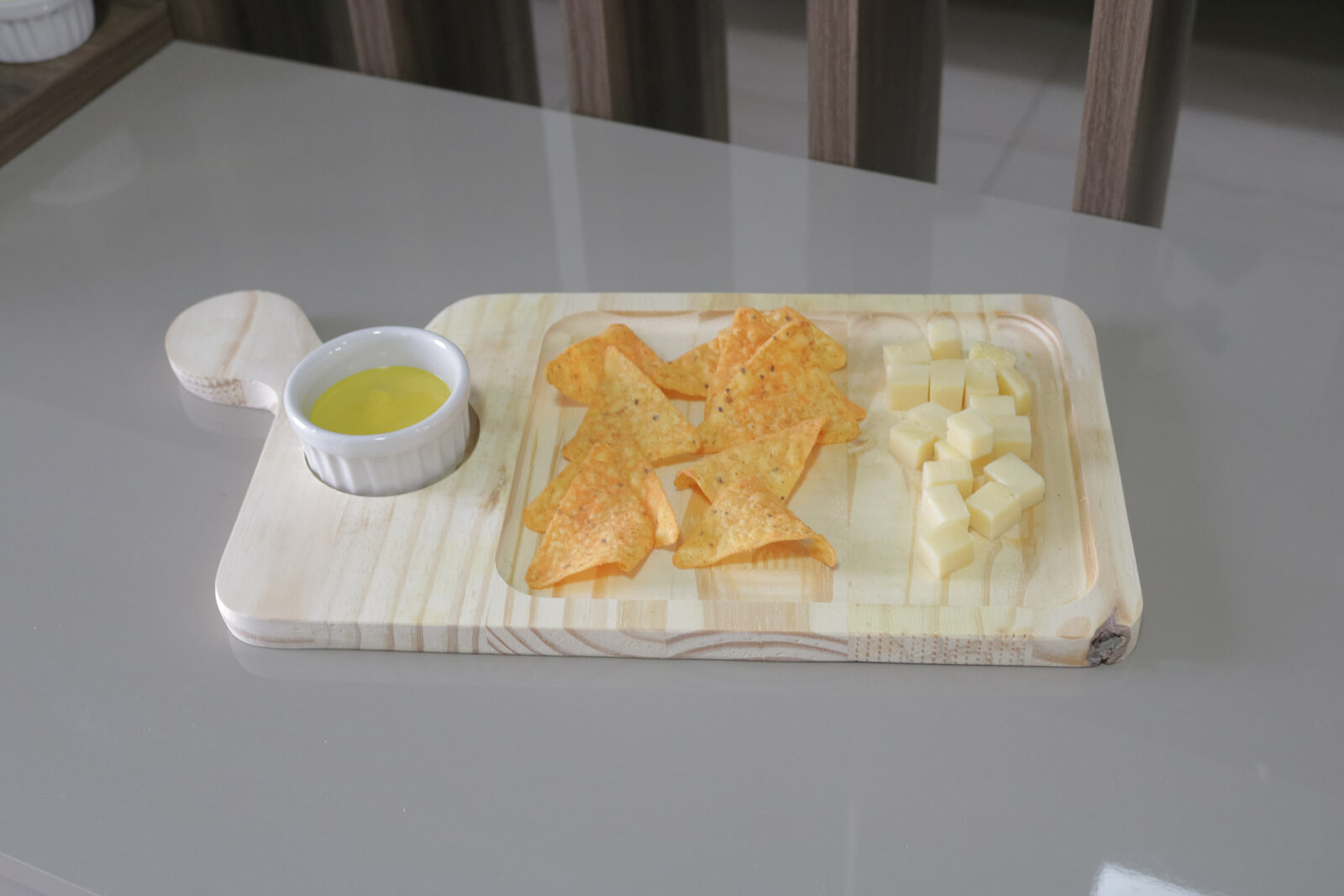 Tábua de madeira clara para petiscos com um ramekim de porcelana, nachos e queijos servidos