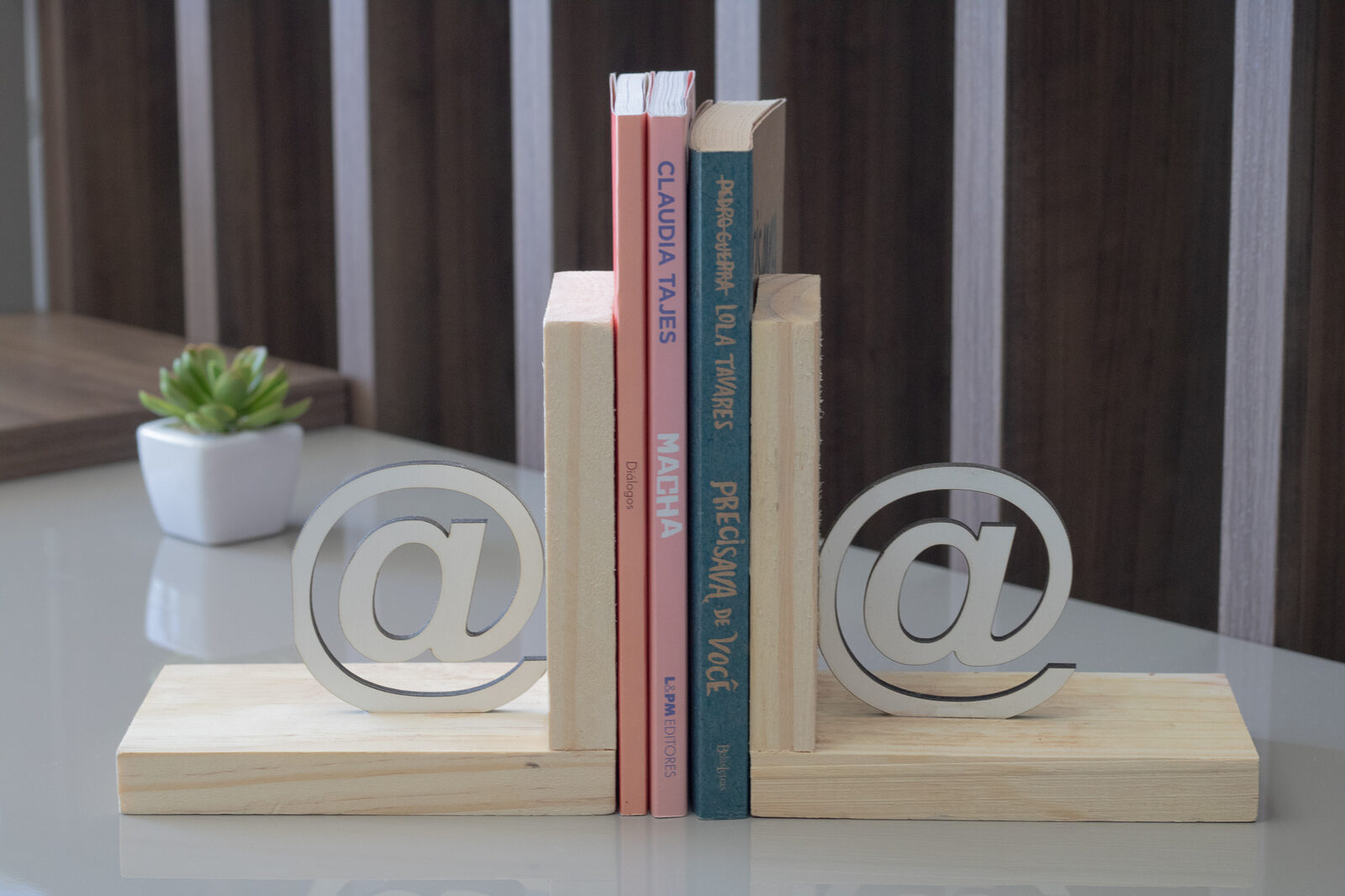 Par de aparadores de livros de madeira pinus com recorte em formato de arroba segurando livros sobre mesa
