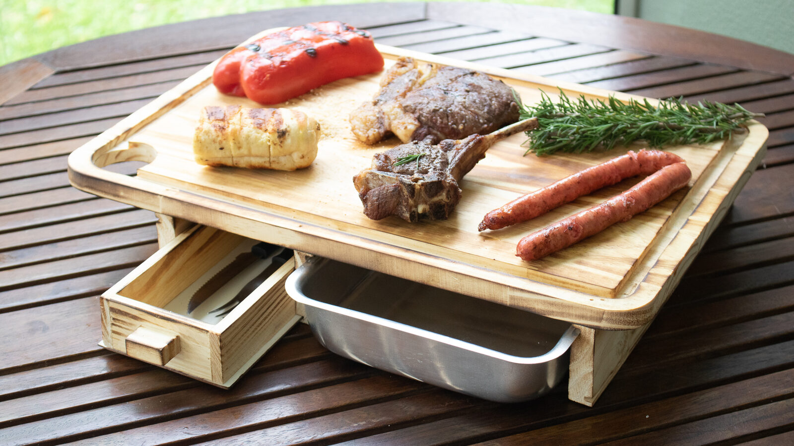 Tábua de churrasco de madeira com bandeja de inox e porta-talher servindo carnes e pães em mesa externa